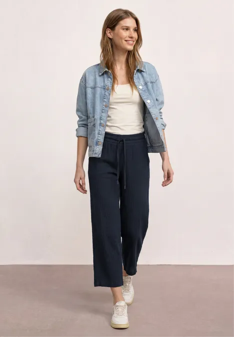 Femme en pantalon large bleu marine, débardeur blanc et veste en jean, baskets blanches sur fond neutre.