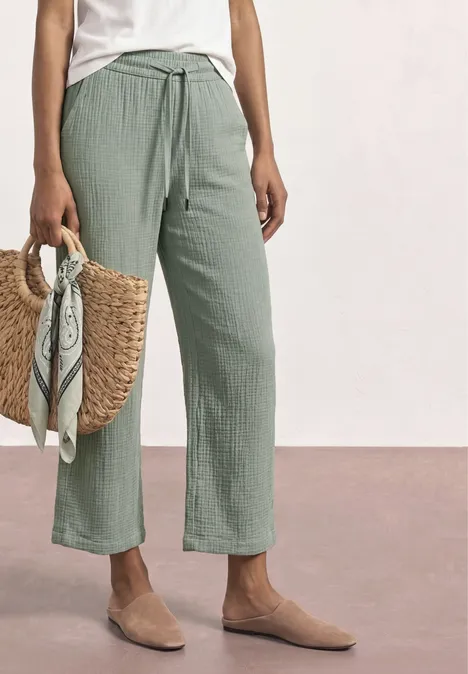 Weite, hellgrüne Leinenhose mit Tunnelzug, dazu neutrale Slipper und geflochtene Strohtasche mit Tuch.