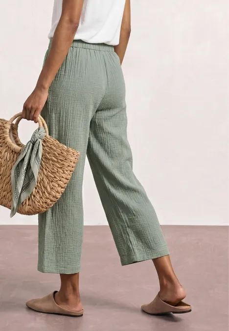 Person in weiten grünen Leinenhosen, weißem Top, mit geflochtenem Korb und flachen Slippern.