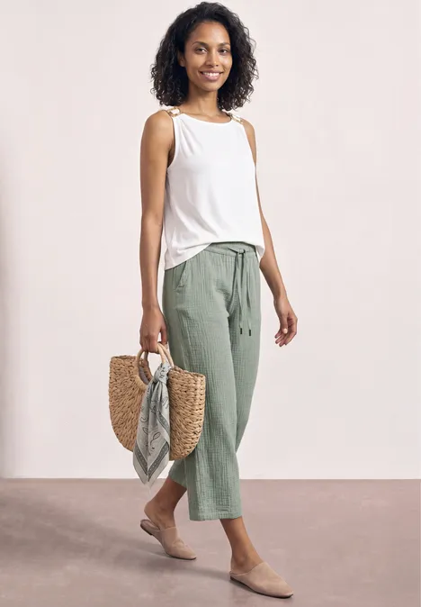 Frau in weißem Top, grünen 7/8-Hosen und Basttasche mit Tuch vor neutralem Hintergrund.