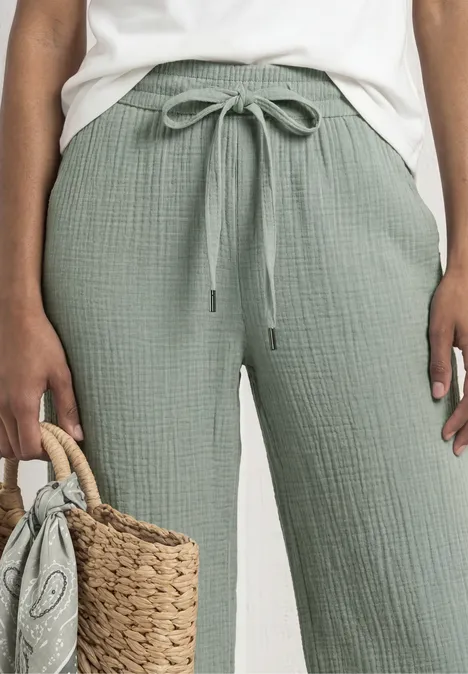 Frau trägt hellgrüne, lockere Baumwollhose mit Kordelzug und hält eine geflochtene Strohtasche.