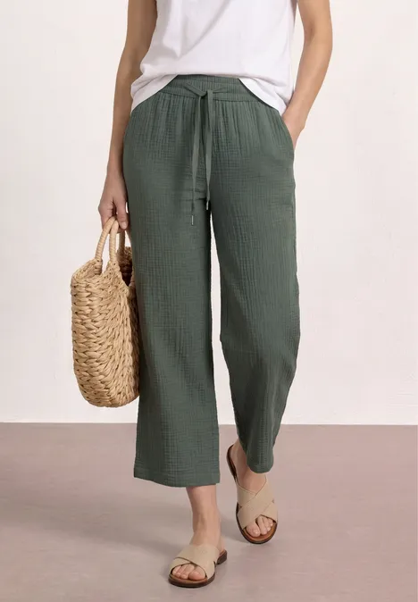 Frau in weißem Top und grüner Leinenhose, trägt Basttasche und Sandalen.