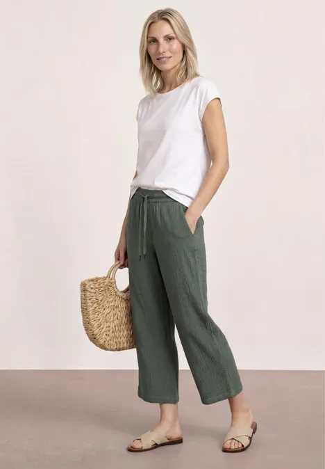 Frau im weißen T-Shirt mit weiten grünen Hosen, Sandalen und Basttasche vor hellem Hintergrund.