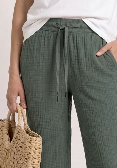 Person trägt weiße Bluse und grüne, lockere Hose mit Kordelzug, hält geflochtenen Strohkorb.