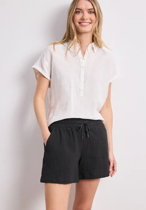 Frau in weißer Bluse und schwarzen Shorts steht lässig mit Händen in den Taschen vor weißem Hintergrund.