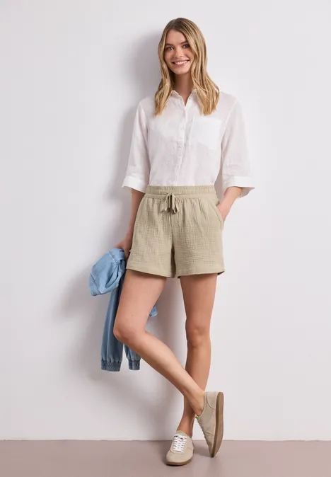 Femme en chemise blanche et short kaki s'appuyant à un mur, tenant une veste en jean à la main.