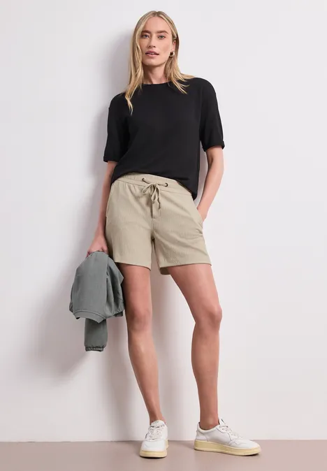 Eine Frau trägt schwarzes T-Shirt, beige Shorts und weiße Sneaker, hält eine Jacke vor weißer Wand.
