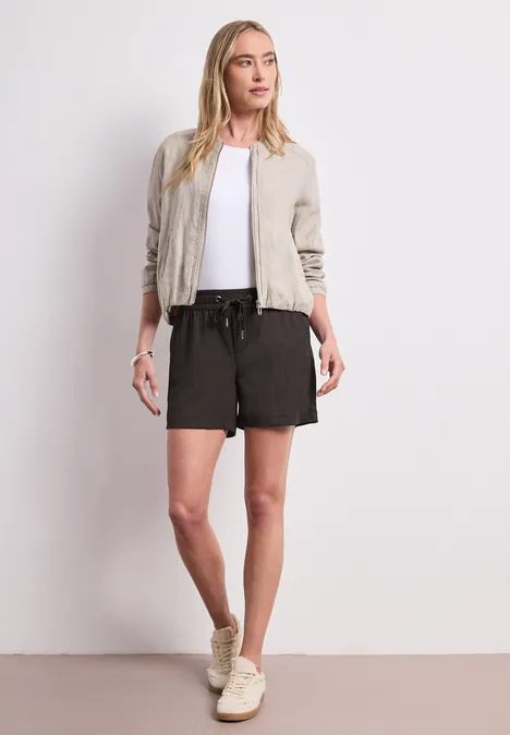 Frau in legerem Outfit mit hellbeiger Jacke, weißem Shirt, schwarzen Shorts und Sneakers.