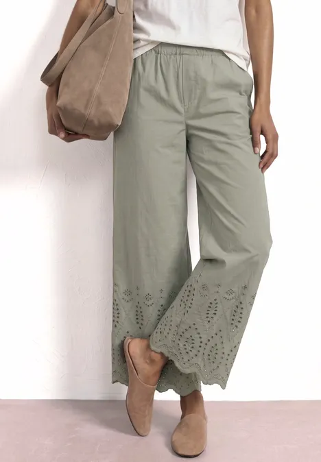 Frau trägt hellgrüne, weit geschnittene 7/8-Hose mit Spitzenbesatz am Saum, weiße Bluse und Mokassins.