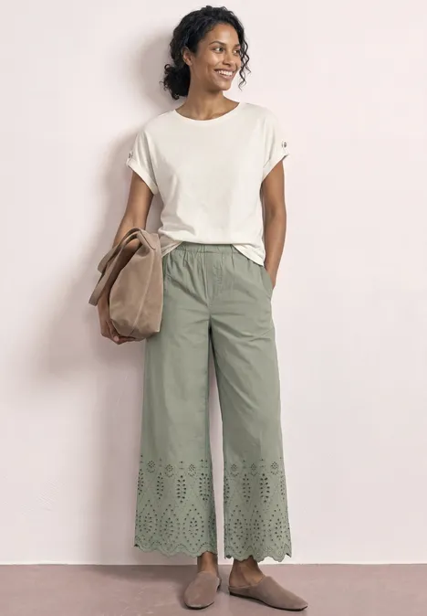 Frau in lockerem weißen T-Shirt, grünen, ausgestellten Hosen mit Lochstickerei am Saum, beigen Schuhen und Tasche.