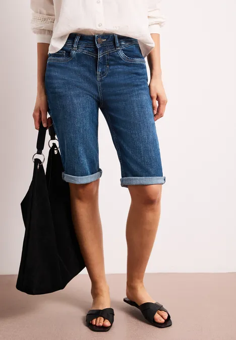 Frau trägt sommerliche Bermudashorts aus Jeans, weiße Bluse, schwarze Sandalen und Tasche.