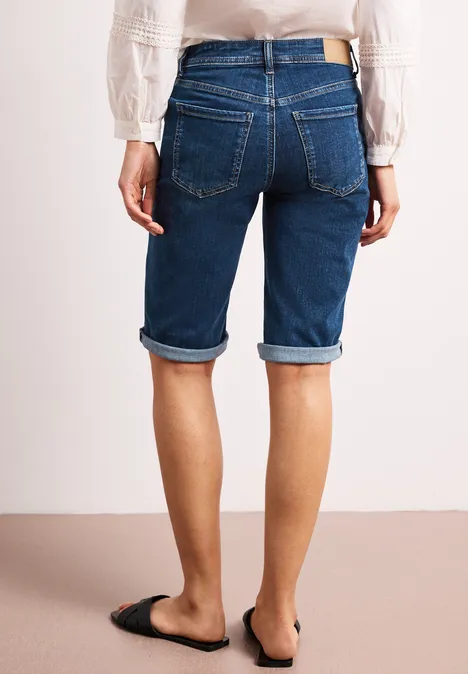 Rückansicht einer Person in knielangen, aufgerollten Jeans-Shorts und schwarzen Slip-On-Schuhen.