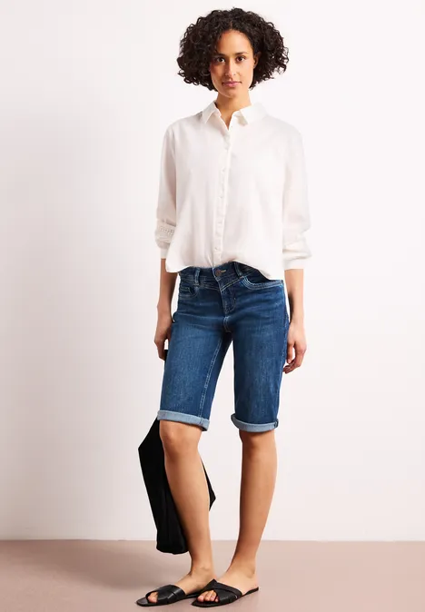 Model trägt weißes Hemd, blaue Jeans-Bermudashorts, schwarze Sandalen und hält eine Tasche.