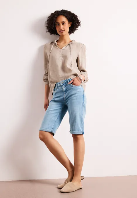 Frau in beiger Leinenbluse und hellblauen Jeans-Knielangen Shorts steht vor weißer Wand in beigen Ballerinas.