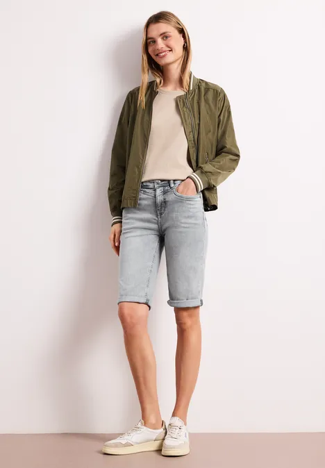 Frau in khaki Blouson, beigem Top, hellblauen Jeans-Bermuda-Shorts und weißen Sneakern vor neutralem Hintergrund.