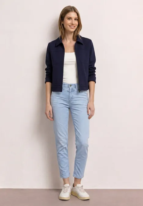 Person steht, trägt dunkelblaue Jacke, weißes Top, helle Jeans und weiße Sneaker.