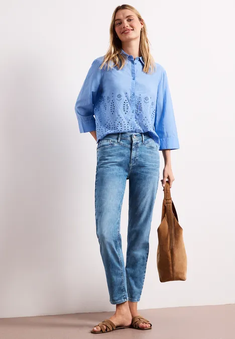 Frau in blauem Spitzenoberteil, Jeans und Sandalen, hält eine braune Tasche.