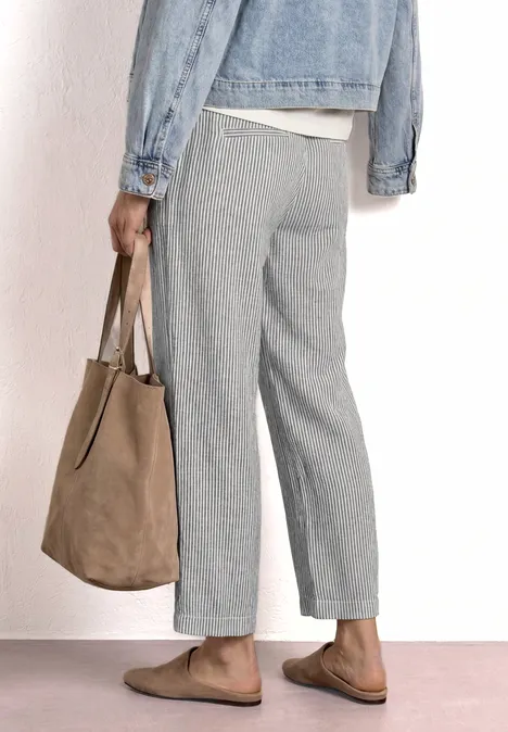 Person vue de profil portant un pantalon gris, une veste en jean, des mocassins bruns et un sac en daim beige.
