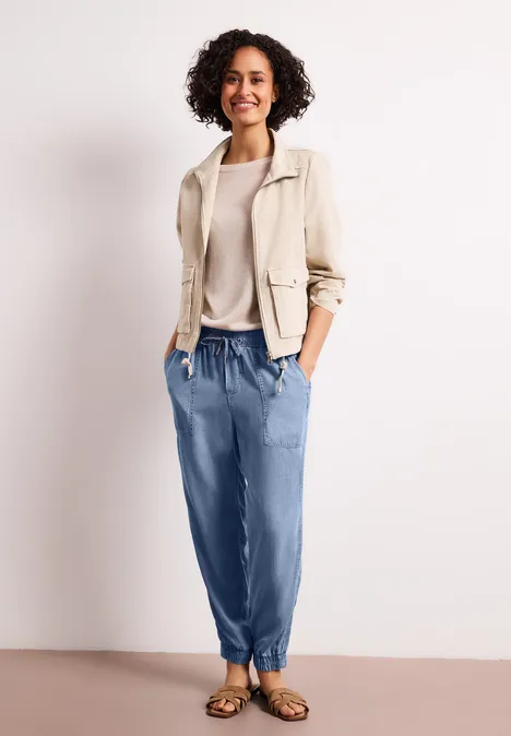 Frau in beigem Blazer, hellem Pullover, blauen Jogging-Jeans und Sandalen mit Händen in den Taschen.