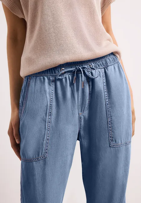 Person in lockeren, hellblauen Jeans mit Gummizug und großen Taschen, dazu beiger Pullover.