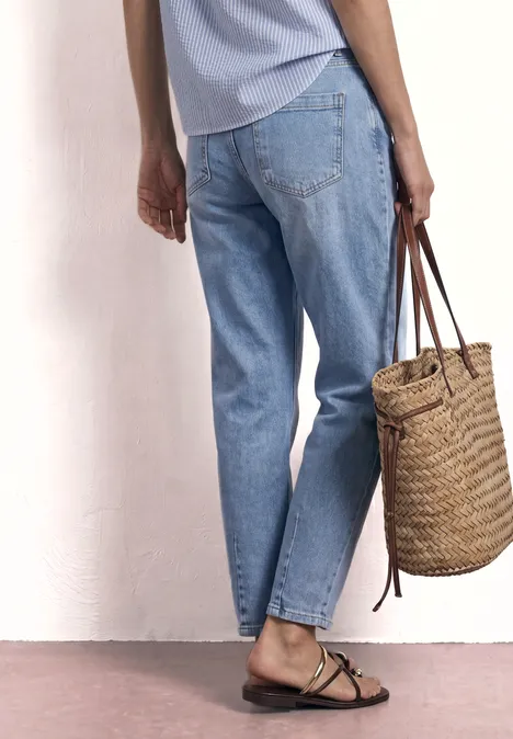 Eine Person in hellen Jeans und Sandalen hält eine geflochtene Strohhandtasche vor einer weißen Wand.