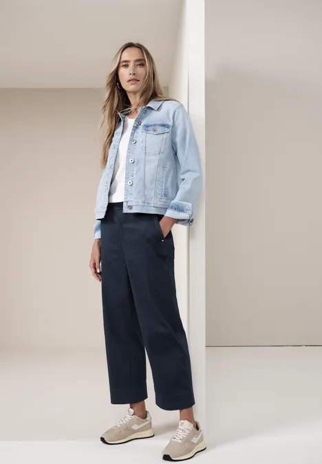 Frau lehnt an Wand, trägt Jeansjacke, weiße Bluse, dunkle weite Hose und Sneakers.