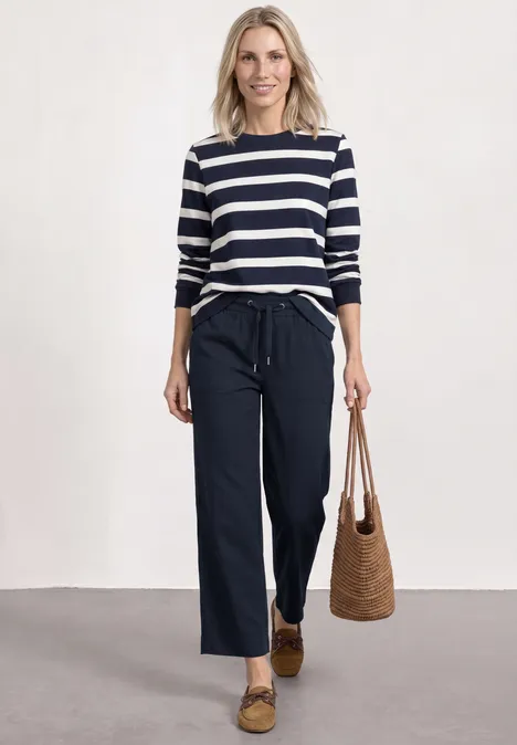 Frau mit blau-weiß gestreiftem Shirt, weiten dunklen Hosen, Basttasche und Loafers.