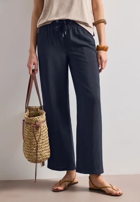 Femme en pantalon noir ample à cordon, tongs et sac en osier tressé.