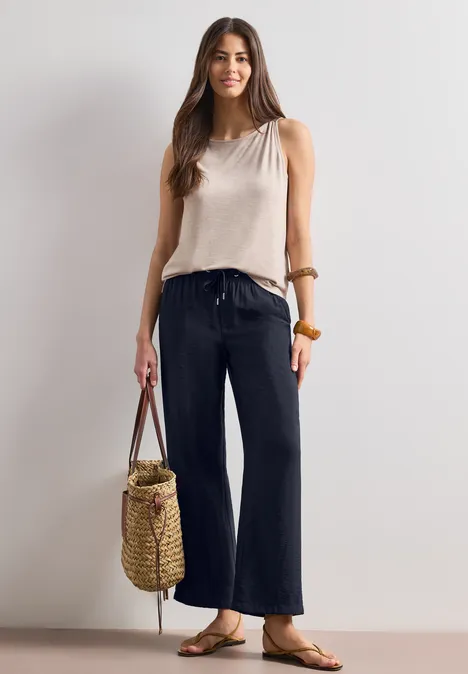 Femme en jean large bleu foncé, top sans manches, sandales et sac en osier.