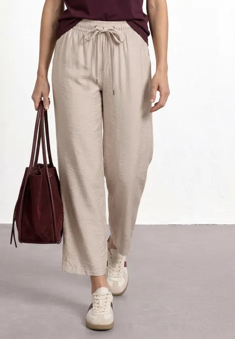 Femme porte un pantalon large en lin beige à taille smockée, baskets blanches et sac marron.