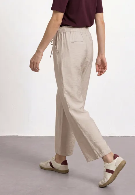 Personne en marche, porte un pantalon beige, des mocassins crème à bride marron et un haut bordeaux.