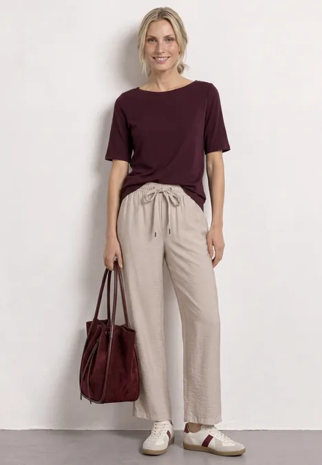 Femme devant un fond blanc, portant un t-shirt bordeaux, un pantalon beige, des baskets blanches et un sac bordeaux.