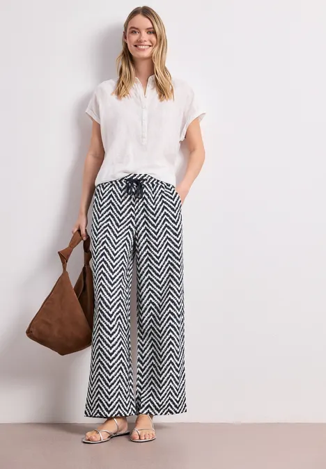 Frau in weißer Bluse und schwarz-weißer Zickzack-Hose mit Schultertasche und Sandalen.