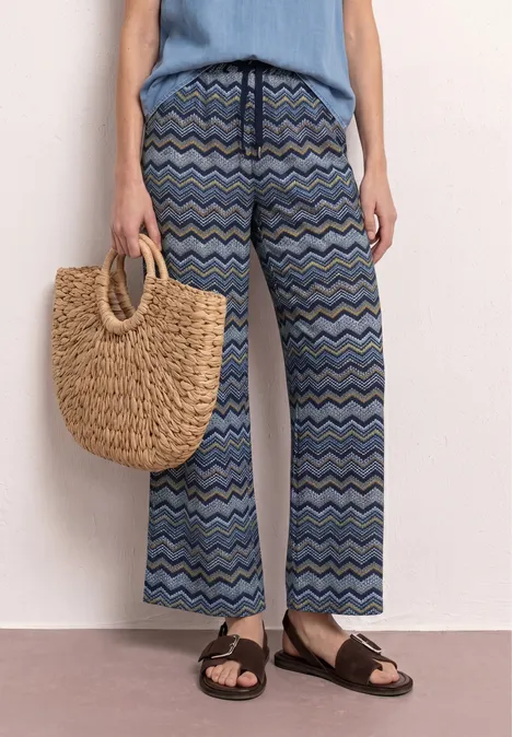 Vrouw met wijd, blauw broek met golvend patroon, nonchalant topje, rieten tas en sandalen.