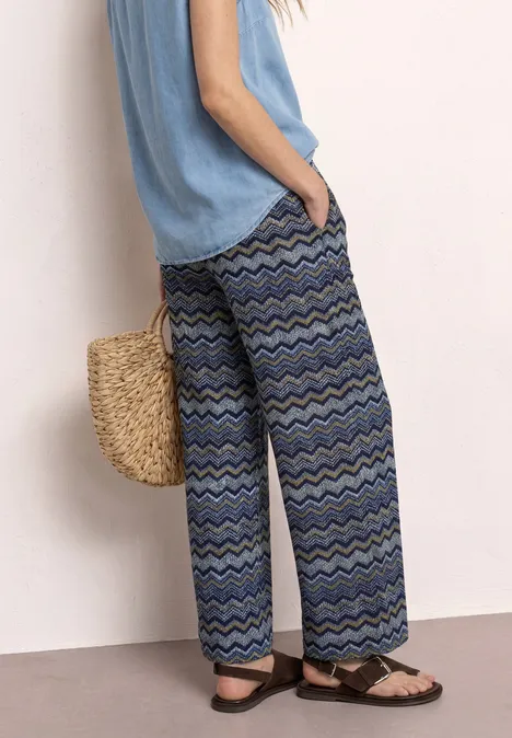 Wijde, blauwe broek met zigzagpatroon, gecombineerd met een luchtig topje, strooien tas en sandalen.