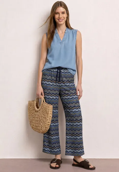 Vrouw met zomerse look: lichtblauw mouwloos topje, wijd broek met patroon, rieten tas en zwarte sandalen.