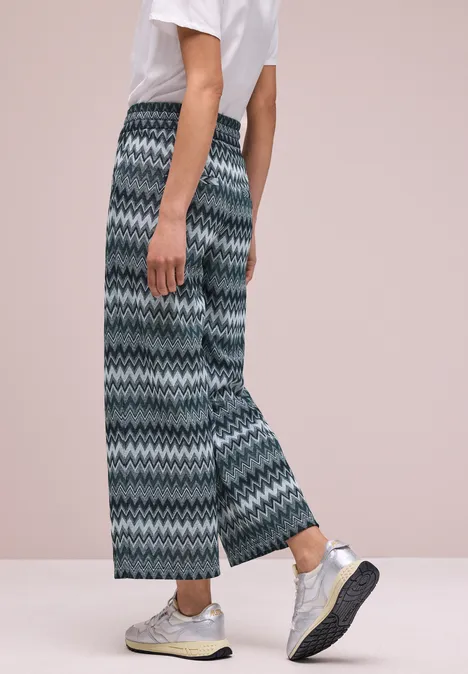 Eine Person trägt weite, gemusterte Hosen, weißes T-Shirt und silberne Sneakers vor hellem Hintergrund.