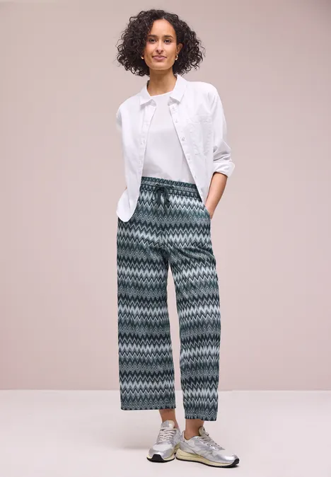 Person steht vor hellem Hintergrund, trägt weiße Bluse über weißem T-Shirt, gemusterte Culotte-Hose und Metallic-Sneaker.