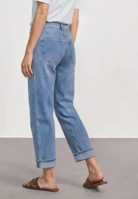Frau in hellblauen, hochgekrempelten Jeans, weißem Oberteil und braunen Sandalen, seitliche Ansicht.