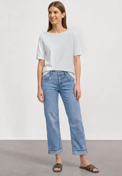 Frau trägt weißes T-Shirt, hellblaue, hochgekrempelte Jeans und braune Sandalen, steht vor neutralem Hintergrund.