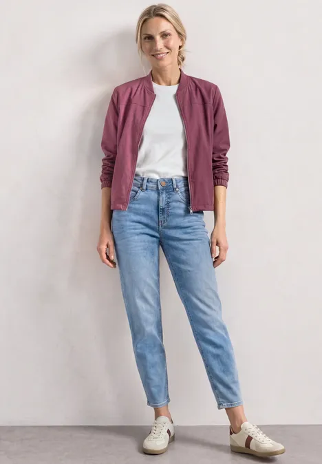 High Waist Barrel Leg Jeans im Loose Fit Light Blue Wash Frau steht in pinker Bomberjacke, weißem Shirt, hellen Jeans und weißen Sneakern vor neutralem Hintergrund.