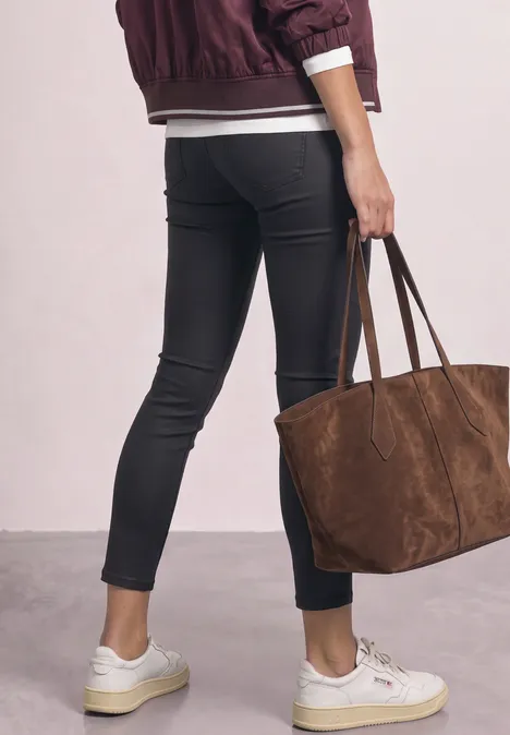 Femme de dos en jean skinny noir, veste bordeaux, baskets blanches, portant un grand sac en daim marron.