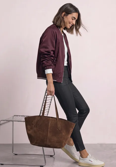 Une femme porte un bomber bordeaux, un jean noir, des baskets blanches et un grand sac en daim marron.