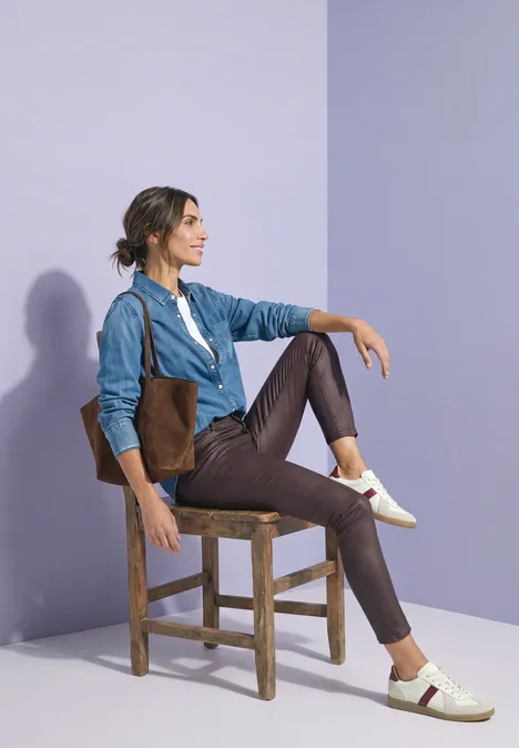 Eine Frau sitzt auf einem Holzstuhl, trägt Jeansjacke, braune Hose und weiße Sneakers vor violettem Hintergrund.