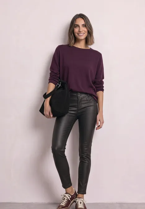 Eine Frau trägt einen lila Pullover, schwarze Lederleggings, Sneakers und hält eine schwarze Tasche vor hellem Hintergrund.