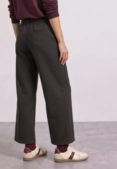 Femme portant un pantalon large noir à rayures fines, chaussettes bordeaux et mocassins blancs sur sol gris.
