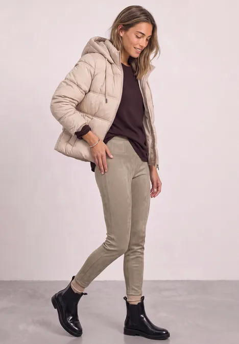 Vrouw in beige gestikte jas, donker shirt, lichte broek en zwarte enkellaarsjes, staand voor een lichte achtergrond.