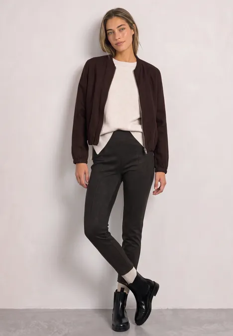 Une jeune femme posant devant un mur clair, veste noire, pull blanc, pantalon et boots noirs.