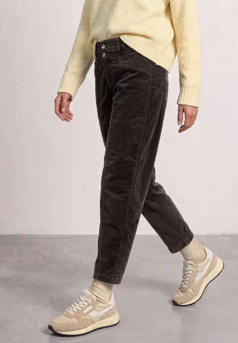 Person trägt dunkelbraune Cordhose, gelben Pulli und beige Sneaker auf grauem Boden