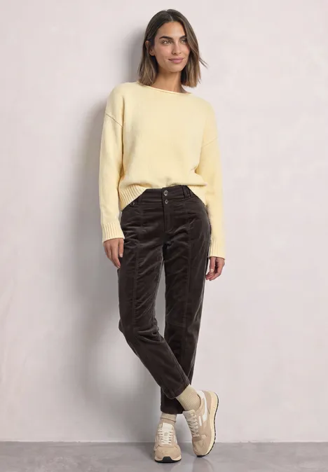 Frau in gelbem Pullover, dunklen Cordhosen und beigen Boots vor hellem Hintergrund.